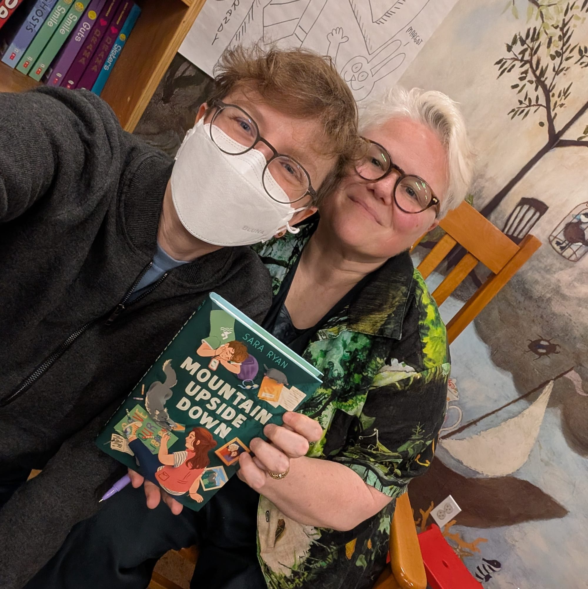 Two white nonbinary people in their 40s and 50s looking super queer and happy, one of them holding a copy of the book.
